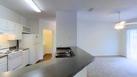 This single-floor residence is thoughtfully arranged to maximize functionality across its compact footprint. The layout, as seen in the 3D floor plan, features an open-concept design that connects the kitchen and living areas seamlessly.

The kitchen serves as a central hub, anchored by a peninsula with a dark countertop surface and a double stainless steel sink. A white refrigerator and additional appliances are integrated into the space, with white cabinetry providing ample storage. A skylight-style flush ceiling fixture illuminates the kitchen area, complementing the textured ceiling finish throughout.

Adjacent to the kitchen is a dedicated laundry area, accessible through a doorway and featuring wood-style flooring that contrasts with the carpeting found in the surrounding rooms. The living areas are finished with neutral-toned carpet, contributing to a cohesive and understated aesthetic throughout.

The floor plan also incorporates a full bathroom with standard fixtures, additional rooms suitable for use as bedrooms or flexible living spaces, and closet storage. The clean white walls and straightforward layout create a bright, practical environment well-suited for everyday living.