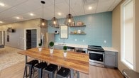 This well-appointed single-floor communal space seamlessly blends a fully equipped kitchen with a relaxed lounge area, creating an inviting environment for gathering and socializing. The kitchen is a standout feature, highlighted by a striking light blue textured tile backsplash that stretches across the full wall, paired with sleek gray cabinetry and white countertops. A substantial live-edge wood communal table anchors the space, surrounded by black industrial-style stools. Overhead, a trio of geometric wire pendant lights adds a contemporary, industrial-chic touch. Stainless steel appliances, including a full-size refrigerator and range, provide full functionality, while rustic floating wood shelves display decorative accents. South Dakota-themed artwork and framed prints lend a regional character throughout. The adjacent lounge area features a generously sized sectional sofa dressed with colorful accent pillows, centered on a patterned area rug. Teal side tables punctuate the neutral, warm-toned walls, while a large window floods the room with natural light and views of the surrounding landscape. Recessed lighting throughout and a drop ceiling ensure a clean, polished finish across both areas.