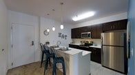 This single-story space showcases a contemporary, open-concept kitchen designed for both functionality and style. The kitchen is anchored by a sleek white peninsula that doubles as a breakfast bar, offering casual seating with industrial-style metal bar stools. Dark espresso-toned cabinetry lines the walls, providing ample storage while creating a striking contrast against the crisp white countertops and light-colored walls. Stainless steel appliances, including a refrigerator, electric range, and over-the-range microwave, lend a polished, modern finish to the space. Three pendant lights hang at varying heights above the breakfast bar, adding a layered lighting element that complements the overhead flush lighting. The kitchen benefits from a bright, well-lit atmosphere courtesy of both the pendant fixtures and a large overhead light panel. The open layout allows the kitchen to flow seamlessly into the surrounding living areas, making it an ideal space for both cooking and entertaining. Neutral flooring and clean wall finishes throughout contribute to a cohesive, contemporary aesthetic.