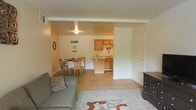 This single-floor unit offers a practical and welcoming open-concept layout that seamlessly connects the main living areas. The living room provides a comfortable space anchored by soft carpeting and a patterned area rug, with easy access to the rest of the unit. A dark wood dresser adds additional storage near the entry area. Adjacent to the living room, the dining area features a wood dining table and chairs set on light hardwood-style flooring, creating a warm and functional space for meals. The kitchen is efficiently designed with honey-toned wooden cabinetry, a light-colored countertop, and a peninsula that offers a natural division between the kitchen and dining areas. The kitchen's layout maximizes functionality while maintaining an open feel to the surrounding spaces. Throughout the unit, the warm beige and neutral wall tones complement the light flooring, contributing to a bright and cohesive ambiance. Ceiling lighting provides consistent illumination across the main living areas. The overall design emphasizes practicality and comfort, making efficient use of the available space while maintaining a welcoming atmosphere throughout.
