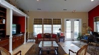 Located at 4545 Cary Glen Blvd in Cary, North Carolina, this single-floor space offers a well-appointed and inviting interior designed for comfort and functionality. The main lounge area immediately draws attention with its soaring vaulted ceilings that lend a sense of openness and grandeur to the room. Warm, neutral-toned walls are complemented by light tile flooring, creating a clean and welcoming atmosphere throughout.

Natural light flows generously into the space through a series of large windows dressed with plaid Roman shades, adding a classic and tailored aesthetic. French doors with glass panels open directly to the outdoors, further enhancing the bright and airy feel of the interior while providing convenient exterior access.

The seating area is anchored by a rich brown leather sofa accented with bold red throw pillows, paired with an ornate wood and glass coffee table set atop a traditional patterned area rug. A decorative wrought-iron accent chair adds an elegant touch to the arrangement. Architectural details such as white decorative columns and pilasters frame the space with a refined, traditional character.

Additional furnishings and cabinetry along the walls provide practical storage and display options, while recessed lighting overhead ensures the space remains well-lit at all hours. Overall, the space combines classic design elements with a warm, functional ambiance.