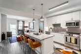 This well-appointed single-floor residence showcases a contemporary design with a cohesive, modern aesthetic throughout. The open-concept kitchen serves as the heart of the home, featuring crisp white cabinetry extending to the ceiling, a textured tile backsplash, stainless steel appliances including a French door refrigerator, gas range, and built-in microwave. A generously sized center island with a quartz countertop and seating provides both additional workspace and a casual dining option, illuminated by stylish pendant lighting overhead. Wood-look flooring ties the kitchen and living areas together seamlessly. The living room offers a comfortable, inviting atmosphere with large windows and a sliding door that opens to a private outdoor space, filling the room with natural light. The primary bedroom is a serene retreat, featuring a deep charcoal accent wall, plush carpeting, and a large window with city views. It connects directly to a spacious walk-in closet with abundant wire shelving and storage. Two well-designed bathrooms are finished with light tile flooring, white floating vanities, quartz countertops, large mirrors, and clean lines. One bathroom includes a glass-enclosed shower, while the other features a shower-tub combination. Throughout the residence, a consistent palette of neutral tones and modern finishes creates an elegant and functional living environment.