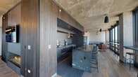 Located at 1353 W Lake St in Chicago's West Loop neighborhood, this single-floor space exemplifies a sophisticated industrial-modern aesthetic. Exposed concrete ceilings and structural columns serve as defining architectural elements throughout, complemented by wide-plank reclaimed hardwood flooring that adds warmth to the raw materiality of the space.

The open-plan layout connects the living and kitchen areas seamlessly. The chef's kitchen is a standout feature, anchored by a dramatic veined marble island with integrated bar seating. Dark, matte cabinetry lines the walls, framing a professional-grade gas range with a stainless steel hood, large-format tile backsplash, and panel-ready refrigeration. Sliding dark wood panels elegantly conceal portions of the kitchen when desired. Warm wood upper cabinetry with under-cabinet lighting adds contrast to the otherwise cool, grey palette.

The living area is expansive and light-filled, with floor-to-ceiling steel-framed windows offering sweeping panoramic views of the Chicago skyline. Exposed concrete columns punctuate the space, while curated seating arrangements and pendant lighting define distinct zones for lounging and conversation. The overall ambiance is refined yet industrial—sophisticated, open, and thoughtfully detailed throughout.