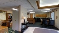 Located at 500 Garden Ln in Huntington, West Virginia, this single-story facility offers a thoughtfully designed interior that balances a professional atmosphere with a homey, inviting feel. The entrance greets visitors with a "Welcome Home to Garden Place" wall inscription, immediately setting a warm and personal tone. The reception area features light tile flooring that transitions into carpeting, a classic console table with a decorative lamp, and a well-appointed front desk surrounded by wood-toned cabinetry. Overhead, a distinctive framed skylight with integrated lighting serves as a notable architectural element, flooding the central area with bright, even illumination alongside recessed drop-ceiling panel lights. The administrative workspace is equipped with filing systems, organized shelving stocked with reference materials, and ergonomic office seating, reflecting a fully functional and organized operations area. Warm beige walls and wood-accented furnishings throughout contribute to a cohesive, comfortable aesthetic. The overall layout is open and accessible, designed to accommodate both staff workflows and the comfort of visitors and residents. The space conveys a professional yet nurturing environment, characteristic of a dedicated care or service facility.