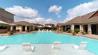 This outdoor pool area offers a resort-style experience designed for relaxation and leisure. The centerpiece is a generously sized swimming pool with crystal-clear, vibrant blue water that immediately evokes a sense of luxury and comfort. A standout feature is the set of in-water lounge chairs positioned at the pool's edge, allowing guests to recline partially submerged — perfect for cooling off while soaking up the sun.

The pool deck is thoughtfully appointed with additional chaise lounges and seating areas arranged along both sides. Patio umbrellas provide shaded retreats, ideal for those seeking relief from direct sunlight. The surrounding hardscape features a well-maintained tiled or stone deck that frames the pool with a clean, polished aesthetic.

Flanking the pool on either side are attractive low-rise structures with hip roofs, likely housing amenity spaces or cabanas, adding architectural charm to the environment. Lush landscaping and potted greenery are integrated throughout, softening the hardscape and adding a natural, welcoming ambiance.

In the background, additional residential buildings are visible, providing context to the community setting. The open sky above, with scattered clouds, enhances the bright, airy atmosphere. Overall, this pool area is a well-designed outdoor amenity that combines functionality with upscale visual appeal.