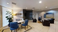 This single-floor space offers a well-designed interior that blends modern aesthetics with functional comfort. The main area serves as both a lounge and meeting space, featuring light-toned ceramic tile flooring throughout that lends the room a bright and clean feel. A defining design choice is the bold charcoal accent wall, which creates a striking focal point and adds depth to the otherwise neutral color palette of white and light gray walls.

The space is furnished thoughtfully with a mix of seating arrangements to accommodate different uses. A white work table surrounded by elegant navy blue upholstered chairs with nailhead trim anchors one side of the room, sitting atop a textured area rug. On the opposite side, a relaxed lounge area features leather-style recliners and an accent armchair in coordinating blue tones, complemented by a cowhide rug and small side tables.

Wall art and decorative mirrors add personality to the space, while recessed ceiling lighting provides even, ambient illumination throughout. A wall-mounted flat-screen display on the accent wall adds a functional media element. The overall ambiance is polished and inviting, making the space well-suited for casual meetings, relaxation, or social gatherings.
