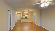 This single-floor residence features an open-concept design that seamlessly connects the living area and kitchen, creating a spacious and functional layout. The living area is finished with warm hardwood-style flooring that extends throughout, complemented by neutral-toned walls that provide a bright and inviting atmosphere. A ceiling fan with a light fixture overhead enhances comfort and air circulation in the main living space.

The kitchen is positioned at the heart of the unit, defined by a peninsula counter that doubles as a breakfast bar, offering both additional prep space and a casual dining option. Warm wood-toned cabinetry lines the kitchen walls, paired with white appliances including a refrigerator and stove. An under-cabinet light fixture ensures the kitchen remains well-lit. A chandelier pendant light hangs above the peninsula, adding a decorative touch.

Near the kitchen, a front entry door provides direct access to the unit, accompanied by a hallway that leads further into the space. A window in the living area allows natural light to filter in, offering a view of surrounding greenery. The overall ambiance is clean, practical, and comfortable.