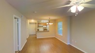 This single-floor residence features an open-concept design that seamlessly connects the living area and kitchen, creating a spacious and functional layout. The living area is finished with warm hardwood-style flooring that extends throughout, complemented by neutral-toned walls that provide a bright and inviting atmosphere. A ceiling fan with a light fixture overhead enhances comfort and air circulation in the main living space.

The kitchen is positioned at the heart of the unit, defined by a peninsula counter that doubles as a breakfast bar, offering both additional prep space and a casual dining option. Warm wood-toned cabinetry lines the kitchen walls, paired with white appliances including a refrigerator and stove. An under-cabinet light fixture ensures the kitchen remains well-lit. A chandelier pendant light hangs above the peninsula, adding a decorative touch.

Near the kitchen, a front entry door provides direct access to the unit, accompanied by a hallway that leads further into the space. A window in the living area allows natural light to filter in, offering a view of surrounding greenery. The overall ambiance is clean, practical, and comfortable.