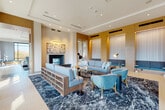 This elegantly designed amenity floor presents a seamless blend of modern sophistication and inviting comfort across a generously proportioned open layout. The grand main lounge anchors the space with soaring ceilings, dramatic dark marble-style columns, and large-scale floor-to-ceiling windows that flood the interior with natural light and frame picturesque outdoor views of the surrounding treeline and cityscape. Light porcelain tile flooring flows throughout, unifying the distinct functional zones.

The primary seating area is appointed with plush powder-blue sofas and velvet accent chairs arranged atop a bold navy abstract area rug, complemented by a sleek fireplace and curated wall art. Gold-accented floor lamps and illuminated wall niches add warm, layered lighting throughout. A separate lounge zone with warm leather chairs and a sculptural stone coffee table offers an intimate retreat beside floor-to-ceiling windows.

A dedicated game room features a billiards table, additional fireplace, and ample lounge seating, while a stylish bar area impresses with a striking blue-tiled island, marble countertops, wood-paneled ceiling, and brass pendant lighting. Elevator access and a well-proportioned transition area with casual dining seating complete this thoughtfully curated communal space.