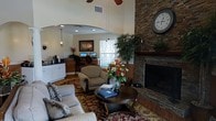 This single-floor space exudes a welcoming, lodge-inspired ambiance that balances comfort with functionality. The focal point of the main lounge area is a floor-to-ceiling stacked stone fireplace, topped with a classic oversized clock and a timber mantel, lending a rustic yet refined character to the room. Soaring vaulted ceilings amplify the sense of openness, while a ceiling fan ensures year-round comfort. The seating arrangement — featuring a plush sofa and upholstered armchairs arranged around a dark wood coffee table atop a richly patterned area rug — invites relaxation and conversation. A vibrant floral centerpiece adds a lively, decorative touch to the lounge. Adjacent to the main seating area, a business or workstation nook is framed by an elegant arched opening, housing multiple computer terminals and cabinetry, making it a practical resource hub. Recessed lighting and wall sconces provide warm, layered illumination throughout. A large indoor plant near the fireplace introduces a refreshing natural element, while natural light filters in through nearby windows, enhancing the overall warmth and livability of the space.
