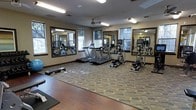 This fitness center is thoughtfully designed to provide a functional and comfortable environment for exercise. The space is outfitted with a selection of modern cardio equipment, including treadmills, an elliptical trainer, and a stair climber, all arranged to allow for ease of movement throughout the room. Large framed mirrors line the walls, enhancing the sense of openness while allowing users to monitor their form during workouts. A step platform and exercise mat are also available, supporting a variety of floor-based and aerobic exercises. The room is finished with a patterned carpet that adds a touch of warmth and visual interest to the space. Natural light filters in through multiple windows, creating a bright and inviting atmosphere, while a ceiling fan provides added airflow and comfort during workouts. Recessed and overhead fluorescent lighting ensures the space remains well-lit at all hours. The neutral-toned walls and dark wood trim detailing along the baseboards and mirror frames contribute to a clean, polished aesthetic. Overall, this fitness center is well-suited to support a range of workout routines in a welcoming and practical setting.