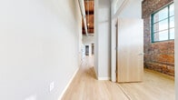 Located at 812 SW Washington St in downtown Peoria, Illinois, this one-bedroom, one-bathroom loft blends industrial character with modern finishes across a single thoughtfully laid-out floor. The space is defined by striking exposed brick walls, soaring ceilings with warm wood-beam detailing, and visible industrial ductwork that reinforces its loft aesthetic. Light hardwood flooring runs throughout, lending a clean, cohesive feel to every room.

A long hallway connects the living areas and leads to the spacious open living room, where oversized factory-style windows — framed by original brick — flood the space with generous natural light and create an airy, open ambiance. The kitchen features white cabinetry, white appliances including a range and refrigerator, and an in-unit washer, all neatly arranged along one wall.

The bedroom is a comfortable, private retreat with its own exposed brick accent wall and a dedicated closet with built-in shelving and hanging rods. The bathroom is clean and functional, offering a full tub-shower combination with crisp white surrounds and wood-look flooring. Two hallways provide smooth circulation throughout the unit, tying together the practical layout with the space's bold, urban industrial character.