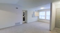 This single-floor residence offers a clean and functional layout designed with everyday comfort in mind. The main living area is finished with plush wall-to-wall carpeting in a neutral tone, creating a warm and inviting atmosphere throughout. Light-colored walls and a smooth ceiling contribute to an airy, open feel that makes the space feel generously sized.

A defining feature of this layout is the open pass-through connecting the living area to the kitchen. The kitchen is equipped with rich, dark cabinetry that provides a striking contrast against the lighter walls, and the pass-through counter offers a convenient breakfast bar or serving area. The kitchen is fully enclosed yet visually connected to the main living space, enhancing the sense of flow and openness.

Large windows along the exterior wall allow abundant natural light to fill the living area, creating a bright and welcoming environment throughout the day. Multiple ceiling vents and floor-level air returns are integrated discreetly into the design, ensuring year-round climate comfort. The neutral color palette and straightforward layout make this space highly versatile and easy to personalize, suited to a wide range of furnishing styles and living preferences.