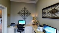 This single-floor space is thoughtfully designed as a dedicated home office, offering a refined and functional environment for work. The room features a built-in L-shaped or wrap-around workstation finished with light granite countertops and crisp white cabinetry below, providing generous workspace and ample storage. The dual-monitor setup, complemented by printers on each side, speaks to a well-planned, productivity-focused layout capable of accommodating multiple users simultaneously. The walls are painted in a soft, muted sage green, lending a calm and focused ambiance to the space. Crown molding along the ceiling adds an elegant architectural touch, elevating the overall aesthetic. Decorative wrought-iron wall art serves as a striking focal point, bringing in an ornate, classic design element that is echoed by a framed piece on the adjacent wall. A warm table lamp paired with a floral arrangement on the countertop adds a welcoming, homey quality to what could otherwise feel like a strictly utilitarian room. The cohesive blend of practical built-ins, tasteful ornamentation, and warm lighting makes this home office both a comfortable and highly efficient workspace.