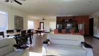 This single-story space is designed as a multifunctional gathering area that seamlessly integrates several distinct zones within an open floor plan. The lounge area is anchored by plush white leather sofas, creating a comfortable seating arrangement suited for socializing or relaxing. Adjacent to the lounge, a pool table serves as a central game room feature, accompanied by bar-height dining seating along the perimeter — ideal for casual meals or social interactions.

The kitchen is well-appointed with dark wood cabinetry, stainless steel appliances including a full-size refrigerator and microwave, and a tiled backsplash that adds visual texture. A raised breakfast bar with dark bar stools defines the boundary between the kitchen and the open living area, while a warm terracotta accent wall adds depth and color contrast to the space.

Natural light filters through multiple windows and a glass door, brightening the neutral-toned walls and tile flooring throughout. A ceiling fan aids in ventilation and comfort. Decorative wall accents and pendant lighting over the kitchen contribute to the space's polished, community-oriented aesthetic.