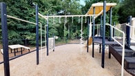 This single-floor space combines a polished interior office environment with an outdoor playground area, making it well-suited for a child-focused or family-oriented business. The interior features a consistent design language throughout, with warm taupe upper walls accented by deep navy-blue wainscoting, white crown molding, and durable light hardwood flooring. Large windows fitted with horizontal blinds and transom glass panels above allow natural light to filter into each room while maintaining privacy.

The office area is equipped with built-in workstations featuring wooden cabinetry, multiple computer monitors, and ergonomic seating, providing a functional workspace for staff. Framed artwork decorates the walls, contributing to a professional yet approachable atmosphere. An adjacent room serves as a utility or print station, housing office equipment and offering access through a glass-paneled interior door. Crown molding is present throughout, reinforcing the space's refined aesthetic.

Outdoors, a well-maintained playground sits on a rubberized safety surface and is framed by lush, mature trees. The play structure includes overhead monkey bars with hanging rings, a spiral climbing pole, a slide, a shaded canopy section, and an interactive activity panel, providing a safe and engaging outdoor environment.