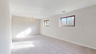 This single-floor space offers a well-organized layout comprising a generously sized living area, multiple bedrooms, a kitchen, and a bathroom. The living area serves as the focal point of the home, finished with light-toned carpeting and neutral walls that create a bright, airy atmosphere. A classic gold chandelier adds a decorative touch overhead, while multiple wood-framed windows along the exterior wall invite abundant natural light and offer views of surrounding greenery.

The bedrooms vary slightly in character: one features blue carpeting and a single window overlooking an outdoor area with mature trees, while others are finished with beige or gray carpet and similarly styled windows that maintain a consistent, understated aesthetic throughout.

The kitchen is equipped with warm oak cabinetry, light countertops, a stainless steel sink, and a gas range with an overhead ventilation hood — providing a practical and functional cooking space. Light vinyl flooring completes the kitchen area.

The bathroom includes an oak vanity with a light countertop, a mirrored medicine cabinet, and light vinyl flooring, maintaining the home's cohesive neutral palette. Overall, the space presents a clean, functional environment with consistent finishes and plentiful natural light throughout.