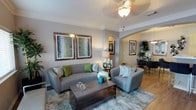 This single-floor residence showcases a thoughtfully designed interior that balances comfort and elegance. The living area serves as the heart of the home, featuring crown molding along the ceiling perimeter and a decorative flush-mount light fixture that provides warm, ambient illumination. Neutral warm-gray walls create a versatile backdrop, complemented by light hardwood-style flooring that extends throughout the open-concept space.

The living room is centered around a plush gray sofa and matching accent chair, accompanied by a walnut-toned coffee table with a lower shelf. Framed botanical artwork adorns the walls, adding visual depth and a touch of nature to the space. An elegant arched passageway connects the living area to the dining space, where pendant lighting and additional framed artwork can be glimpsed, reinforcing the cohesive design throughout.

The dining area, visible through the archway, features dark furniture and lush indoor greenery, adding contrast and vibrancy. Overall, the space exudes a refined yet approachable atmosphere, with architectural details and carefully chosen finishes contributing to a polished, welcoming environment.