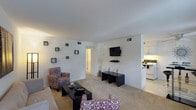 This single-floor residence offers an open and well-organized layout that seamlessly connects the living area to the kitchen. The living room is finished with neutral-toned carpet and bright white walls, creating a clean and inviting atmosphere. A wall-mounted flat-screen television is positioned above a low-profile media shelf, serving as a natural focal point for the seating area. The space is furnished with patterned accent chairs and a sleek black coffee table, complemented by a matching set of side tables that add a cohesive, contemporary touch. A cluster of small square wall frames arranged decoratively on one wall adds visual interest and a personalized aesthetic. Recessed HVAC vents are integrated into the walls, maintaining the streamlined appearance of the room. Adjacent to the living area, the kitchen is visible through an open passageway and features white cabinetry, white appliances, and under-cabinet lighting that provides a bright workspace. A hallway with warm lighting leads further into the unit, suggesting additional rooms beyond the main living space. Overall, the space presents a functional, modern layout with a clean and neutral design palette.
