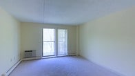 This single-floor space features a carpeted main living area finished in neutral tones, creating a clean and understated atmosphere. The room is characterized by its sliding glass door, which allows natural light to filter in and provides a view of surrounding trees, suggesting a connection to the outdoors. Vertical blinds offer adjustable light control and privacy. Along the walls, baseboard heating units ensure year-round comfort, while a wall-mounted air conditioning unit provides additional climate control. The textured ceiling and light yellow walls contribute to a simple, functional aesthetic. The open layout of the main living area offers flexibility for a variety of furniture arrangements, making efficient use of the available space.