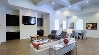 This single-story space showcases a thoughtfully designed living area that combines elegance with functionality. The room's most striking architectural feature is its coffered ceiling, which adds depth and visual interest to the generous vertical space above. Recessed lighting is embedded throughout, complementing the natural light that streams in through multiple arched and rectangular windows fitted with white blinds.

The flooring is finished in warm hardwood tones, providing a cohesive foundation throughout the open layout. A wall-mounted flat-screen television anchors one side of the room, flanked by dark-finished cabinetry that offers both storage and a bold contrast to the bright white walls. Seating is arranged thoughtfully, featuring a light neutral sofa accented with red throw pillows, paired with two upholstered accent chairs in charcoal tones. A glass-topped coffee table and a sleek console table with decorative accents complete the living arrangement. A table lamp with a classic white shade adds warm ambient lighting to the seating area. The overall ambiance is clean, modern, and inviting, with a balanced mix of neutral tones and rich accent colors creating a polished yet comfortable atmosphere.
