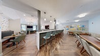 This thoughtfully designed single-floor community clubhouse offers a welcoming and versatile gathering space highlighted by consistent herringbone-patterned hardwood flooring throughout. The open layout flows seamlessly between distinct zones, each with its own character and function.

The central lounge area is anchored by a striking sputnik chandelier suspended from a vaulted ceiling, casting warm light over an arrangement of mid-century modern seating, including plush sofas, accent chairs, and a wall-mounted television framed by large windows that invite natural light. Adjacent to this space, a long bar-style counter with a white marble top and dark paneled base accommodates numerous teal upholstered barstools beneath pendant lighting, creating an ideal social hub.

The kitchenette is well-appointed with grey cabinetry, quartz countertops, a stainless steel refrigerator, built-in oven and warming drawer, a tile backsplash, open wood shelving, and a dedicated coffee station. A smaller nook nearby features a high-top bistro table with wall-mounted TV, perfect for casual conversation or individual use.

Throughout the space, curated artwork, wainscoting, and soft neutral tones create a refined yet approachable ambiance. Double glass-panel entry doors connect the interior to an outdoor covered area, further enhancing the sense of openness and accessibility.