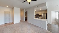This single-floor residence offers a well-appointed, open layout that seamlessly connects the living and kitchen areas. The living room features neutral-toned carpeting, a ceiling fan with light fixture, and French doors that invite natural light while providing access to an outdoor space. The open-concept kitchen is equipped with stainless steel appliances, including a range, over-the-range microwave, and refrigerator, complemented by light-toned cabinetry, a dark tile backsplash, and stone-finish countertops. A peninsula countertop with a built-in sink extends into the living area, making it ideal for casual dining or entertaining. The bedroom is generously proportioned with plush carpeting, a ceiling fan, and its own set of French doors offering additional outdoor access and natural light. The bathroom features a long vanity with dual vessel sinks, gray cabinetry, light stone countertops, and large mirrors, all set atop rich dark hardwood-style flooring. Recessed lighting adds warmth throughout. The overall aesthetic is clean and contemporary, with a neutral color palette and modern finishes that create a bright, cohesive living environment.