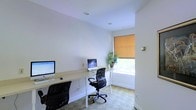 This single-floor space is designed as a functional and efficient workspace. The room features crisp white walls and a smooth white ceiling with recessed lighting, creating a bright and clean atmosphere. A long, light-wood desk runs along the wall, providing ample surface area to accommodate two workstations, each equipped with desktop monitors and ergonomic mesh office chairs. The layout encourages a shared yet focused work environment. A window fitted with a warm-toned roller shade allows natural light to filter into the space while maintaining privacy. A potted green plant positioned near the window adds a touch of nature and visual warmth to the otherwise minimalist setting. The flooring features a patterned carpet that subtly adds texture to the room. The overall design is straightforward and practical, with a neutral color palette that promotes concentration. The recessed ceiling fixtures provide consistent overhead illumination, supplementing the natural light from the window. This workspace is well-suited for collaborative or individual use, offering a calm and organized environment for daily productivity.