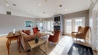 This single-floor community amenity space is thoughtfully designed to serve as a social hub, offering a variety of functional areas within an open, interconnected layout. The main lounge area sets a warm and inviting tone, featuring a classic fireplace framed by a blue accent wall and a wall-mounted television. Comfortable seating — including upholstered sofas, wingback chairs, and accent armchairs — is arranged around a glass-top coffee table, creating an ideal setting for relaxed conversation. Natural light pours in through multiple French doors and large windows, enhancing the bright, airy ambiance throughout. Light hardwood flooring runs continuously across the space, tying each area together cohesively. Adjacent to the lounge, a generously sized dining area accommodates multiple round tables with ornate wrought-iron and upholstered chairs, providing ample space for group dining and events. The kitchen features a wraparound counter with bar-height seating, warm wood cabinetry, stainless steel appliances, and a built-in sink — well-equipped for catering and communal use. Horizontal clerestory windows above the counter allow natural light in while maintaining privacy. A separate fitness room, visible through an open doorway, houses cardio equipment and benefits from its own access to natural light via French doors. Neutral gray walls and recessed lighting throughout lend the space a clean, polished finish.