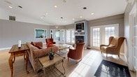 This single-floor community amenity space is thoughtfully designed to serve as a social hub, offering a variety of functional areas within an open, interconnected layout. The main lounge area sets a warm and inviting tone, featuring a classic fireplace framed by a blue accent wall and a wall-mounted television. Comfortable seating — including upholstered sofas, wingback chairs, and accent armchairs — is arranged around a glass-top coffee table, creating an ideal setting for relaxed conversation. Natural light pours in through multiple French doors and large windows, enhancing the bright, airy ambiance throughout. Light hardwood flooring runs continuously across the space, tying each area together cohesively. Adjacent to the lounge, a generously sized dining area accommodates multiple round tables with ornate wrought-iron and upholstered chairs, providing ample space for group dining and events. The kitchen features a wraparound counter with bar-height seating, warm wood cabinetry, stainless steel appliances, and a built-in sink — well-equipped for catering and communal use. Horizontal clerestory windows above the counter allow natural light in while maintaining privacy. A separate fitness room, visible through an open doorway, houses cardio equipment and benefits from its own access to natural light via French doors. Neutral gray walls and recessed lighting throughout lend the space a clean, polished finish.