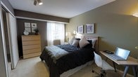 This single-floor residence offers a thoughtfully designed living space with a warm and inviting atmosphere. The bedroom serves as a central highlight, featuring a combination of earthy tones with sage green and deep brown accent walls that create a calming, grounded ambiance. Soft, neutral carpeting underfoot adds to the comfort of the room. A generously sized bed dressed in dark, textured bedding with layered pillows anchors the space, complemented by a warm wooden headboard and a light wood dresser that adds natural character to the room. Framed wall art above the headboard contributes an understated elegance to the décor. Natural light filters in through a window fitted with white sheer curtains and blinds, brightening the space while maintaining privacy. A bedside lamp adds a soft, warm glow for evening ambiance. A compact desk area with a modern white chair is neatly tucked along one wall, making the room both restful and functional for work or study. The overall design blends contemporary and transitional styles, creating a space that feels both practical and aesthetically cohesive.