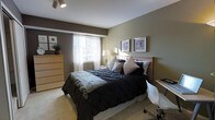 This single-floor residence offers a thoughtfully designed living space with a warm and inviting atmosphere. The bedroom serves as a central highlight, featuring a combination of earthy tones with sage green and deep brown accent walls that create a calming, grounded ambiance. Soft, neutral carpeting underfoot adds to the comfort of the room. A generously sized bed dressed in dark, textured bedding with layered pillows anchors the space, complemented by a warm wooden headboard and a light wood dresser that adds natural character to the room. Framed wall art above the headboard contributes an understated elegance to the décor. Natural light filters in through a window fitted with white sheer curtains and blinds, brightening the space while maintaining privacy. A bedside lamp adds a soft, warm glow for evening ambiance. A compact desk area with a modern white chair is neatly tucked along one wall, making the room both restful and functional for work or study. The overall design blends contemporary and transitional styles, creating a space that feels both practical and aesthetically cohesive.