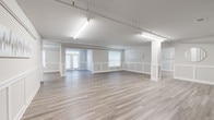 This single-floor commercial space offers a clean, modern interior defined by an open and versatile layout. The main area is expansive and unobstructed, with wide-plank light hardwood flooring running throughout, providing a cohesive and polished aesthetic across the entire space. Crisp white wainscoting lines the lower half of the walls in every room, adding a refined architectural detail that elevates the overall design.

The space is organized into a large open main area that flows into several connected secondary rooms, each benefiting from natural light through double-hung windows fitted with horizontal blinds. The neutral gray-white wall palette keeps the atmosphere bright and adaptable to a wide range of uses.

Overhead, a combination of fluorescent tube lighting and adjustable track lighting systems provides ample, evenly distributed illumination throughout, making the space well-suited for professional or creative uses. Exposed conduit along the ceiling adds a subtle industrial element that contrasts tastefully with the otherwise polished interior finishes.

Structural columns punctuate the main area, defining zones without fully enclosing them, while interior doorways connect the various rooms seamlessly. The floor plan, visible in the dollhouse view, reveals a thoughtfully arranged layout with clear circulation paths and distinct zones that offer both open collaboration areas and more private enclosed rooms.