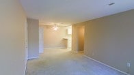 This single-floor residence offers a straightforward and functional layout designed for comfortable everyday living. The main living area is spacious and open, finished with wall-to-wall carpeting in a neutral tone that lends warmth to the space. Soft beige walls reflect light throughout, creating a bright and welcoming atmosphere. A ceiling fan is centrally mounted overhead, providing both airflow and a modest focal point to the room.

The living area flows seamlessly toward a kitchen space, visible through an open passageway. The kitchen features light wood cabinetry and a countertop peninsula or breakfast bar, offering a practical transition between cooking and living areas. The layout encourages an open-concept feel, allowing sightlines to carry from one end of the space to the other.

Adjacent to the main living area, a doorway leads to additional rooms, suggesting a well-connected floor plan. White trim and baseboards throughout provide clean contrast against the beige walls, giving the interior a tidy, cohesive appearance. The overall ambiance is calm and neutral, making the space a versatile canvas well-suited for a variety of interior styles and furnishing preferences.