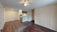 This single-floor unit offers a practical and well-organized layout designed for comfortable everyday living. The open living area serves as the heart of the home, featuring rich, dark hardwood-style flooring, a ceiling fan with light fixture, and a large sliding glass door dressed with vertical blinds that fills the space with natural light and provides access to an outdoor area. The living space flows directly into the kitchen, which is configured in a galley style with a pass-through counter. The kitchen is equipped with warm wood-toned cabinetry offering ample upper and lower storage, a double-basin stainless steel sink, a coil-top electric range, a dishwasher, and a full-size refrigerator, all beneath bright overhead fluorescent lighting and a textured ceiling. The primary bedroom is generously sized, continuing the dark hardwood-style flooring, and benefits from a ceiling fan and natural light from a window. The secondary bedroom features plush carpeting, a window with blinds, and a textured ceiling. The bathrooms include a full bath with a tiled tub-and-shower combination, a louvered closet door for storage, and clean white fixtures. Throughout the unit, neutral wall tones and textured ceilings contribute to a cohesive, understated aesthetic.