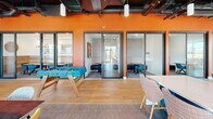 Located at 414 Front St in Santa Cruz, CA, this single-floor amenity and coworking space offers a generously sized, open-plan layout designed to balance productivity with social interaction. The interior showcases a striking geometric coffered ceiling in the lounge zone, contrasted by exposed industrial ductwork and high ceilings throughout the remainder of the floor, creating a dynamic architectural character. Light oak hardwood flooring runs throughout most of the space, complemented by patterned area rugs that define distinct zones.

The lounge area features plush terracotta-hued velvet sofas, an eclectic mix of accent chairs, and a wall-mounted display screen, all bathed in abundant natural light from expansive black-framed industrial-style windows. Adjacent to the lounge, a sleek communal kitchen bar with white quartz countertops and leather bar seating provides a social gathering point beneath a sculptural ring chandelier.

A dedicated game room includes a foosball table and a pool table, anchored by a bold orange accent wall. Glass-enclosed private phone booths and quiet meeting rooms line one corridor, offering focused workspaces. Built-in bench seating with direct access to an outdoor area rounds out this vibrant, multifunctional environment.