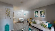 This thoughtfully designed single-floor residence offers a cohesive, contemporary aesthetic highlighted by a neutral gray color palette accented with teal and gold tones throughout. The open-concept living and dining area serves as the heart of the unit, featuring wood-look laminate flooring, a textured ceiling, and a bright front entry door flanked by sidelight windows fitted with blinds, allowing natural light to filter into the space. The living area accommodates a comfortable seating arrangement anchored by a glass coffee table and a patterned area rug, while framed botanical artwork adds visual interest to the walls.

Adjacent to the living area, a defined dining nook provides space for a round white table and chairs, decorated with framed artwork in a coordinated style. The galley-style kitchen features white cabinetry, light countertops, white appliances including a refrigerator and electric range, and a charming arched pass-through that connects to the living space. Wood-look flooring continues through this area seamlessly.

The bedroom is carpeted and generously proportioned, offering a ceiling fan with light fixture, a large window dressed with sheer curtains for soft natural light, and a closet with built-in shelving. Direct access to a private bathroom is conveniently located within the bedroom corridor, alongside an additional closet, providing practical and well-organized storage throughout.