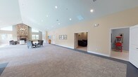 This single-floor community clubhouse offers a diverse range of amenities designed to support both active and social lifestyles. At its heart is an impressive lounge area anchored by a soaring floor-to-ceiling stone fireplace, complemented by rich hardwood flooring, vaulted ceilings with clerestory windows, and plentiful natural light. Comfortable seating arrangements and a tasteful chandelier create a warm, inviting atmosphere ideal for gatherings or relaxation.

The expansive multipurpose hall features soft patterned carpeting, warm yellow-toned walls, and vaulted ceilings with recessed lighting. The open layout accommodates a variety of uses, from community events to casual meetings, with flexible seating and access to an adjacent media or lounge room. Adirondack rocking chairs near sun-filled windows add a welcoming, casual touch.

The kitchenette is finished with crisp white cabinetry, granite countertops, stainless steel appliances, and soft blue walls accented by decorative wall art — a practical and charming space for catering or casual refreshment.

The fitness center is well-equipped with cardio machines, a cable weight system, free weights, and a stability ball, all within a carpeted, recessed-lit room. An additional light-filled studio room with hardwood floors and a pool view provides space for yoga or stretching. Together, these areas form a comprehensive and thoughtfully designed community facility.