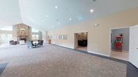 This single-floor community clubhouse offers a diverse range of amenities designed to support both active and social lifestyles. At its heart is an impressive lounge area anchored by a soaring floor-to-ceiling stone fireplace, complemented by rich hardwood flooring, vaulted ceilings with clerestory windows, and plentiful natural light. Comfortable seating arrangements and a tasteful chandelier create a warm, inviting atmosphere ideal for gatherings or relaxation.

The expansive multipurpose hall features soft patterned carpeting, warm yellow-toned walls, and vaulted ceilings with recessed lighting. The open layout accommodates a variety of uses, from community events to casual meetings, with flexible seating and access to an adjacent media or lounge room. Adirondack rocking chairs near sun-filled windows add a welcoming, casual touch.

The kitchenette is finished with crisp white cabinetry, granite countertops, stainless steel appliances, and soft blue walls accented by decorative wall art — a practical and charming space for catering or casual refreshment.

The fitness center is well-equipped with cardio machines, a cable weight system, free weights, and a stability ball, all within a carpeted, recessed-lit room. An additional light-filled studio room with hardwood floors and a pool view provides space for yoga or stretching. Together, these areas form a comprehensive and thoughtfully designed community facility.