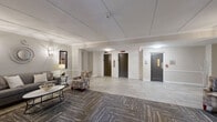 Located at 313 Creek Dr in Wayne, PA, this thoughtfully designed hallway and lobby space offers a gracious and welcoming atmosphere that sets the tone for the building. The layout features a generously sized central lounge area that transitions seamlessly into the main corridor, creating a fluid and functional flow throughout.

The lounge is anchored by a striking stacked-stone fireplace with a rich wood mantel, complemented by elegant sconce lighting and curated artwork on the walls. Comfortable seating, including upholstered armchairs and a plush sofa arranged around a sleek coffee table, adds a residential warmth to the shared space. The flooring combines large-format light stone-look tile with patterned carpet in the seating areas, adding visual texture and defining zones within the open layout.

Crown molding and wainscoting line the walls, lending a classic, refined aesthetic. Two elevator bays framed by a stacked-stone accent wall provide convenient access throughout the building, while a stairwell door is positioned nearby. At the far end, a set of glass-paneled entry doors floods the space with natural light, flanked by symmetrically placed armchairs and framed artwork. The overall ambiance is polished and inviting, reflecting a well-appointed building common area designed for both functionality and comfort.