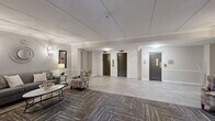 Located at 313 Creek Dr in Wayne, PA, this thoughtfully designed hallway and lobby space offers a gracious and welcoming atmosphere that sets the tone for the building. The layout features a generously sized central lounge area that transitions seamlessly into the main corridor, creating a fluid and functional flow throughout.

The lounge is anchored by a striking stacked-stone fireplace with a rich wood mantel, complemented by elegant sconce lighting and curated artwork on the walls. Comfortable seating, including upholstered armchairs and a plush sofa arranged around a sleek coffee table, adds a residential warmth to the shared space. The flooring combines large-format light stone-look tile with patterned carpet in the seating areas, adding visual texture and defining zones within the open layout.

Crown molding and wainscoting line the walls, lending a classic, refined aesthetic. Two elevator bays framed by a stacked-stone accent wall provide convenient access throughout the building, while a stairwell door is positioned nearby. At the far end, a set of glass-paneled entry doors floods the space with natural light, flanked by symmetrically placed armchairs and framed artwork. The overall ambiance is polished and inviting, reflecting a well-appointed building common area designed for both functionality and comfort.