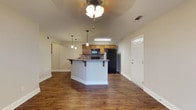 This single-story space showcases an open-concept design that seamlessly connects the living and kitchen areas, creating a sense of spaciousness and easy flow throughout. Rich, dark hardwood-style flooring runs continuously across the entire space, lending warmth and a cohesive aesthetic to the interior.

The kitchen is a functional centerpiece, equipped with medium-toned wood cabinetry providing ample storage. Black appliances, including a refrigerator and microwave, offer a sleek contrast against the cabinetry. A kitchen island with a dark countertop and an overhang for bar seating serves as both a prep space and a casual dining area, bridging the kitchen and living zone.

Lighting is thoughtfully layered throughout the space. Pendant lights hang above the island, a ceiling fan with a light fixture illuminates the living area, and a fluorescent fixture brightens the kitchen workspace. Vaulted or elevated ceilings contribute to the open, airy feel of the interior.

White baseboards and trim provide clean contrast against the flooring, while neutral wall tones keep the space feeling bright. A white paneled entry door and visible HVAC vents complete this well-appointed, move-in-ready layout.