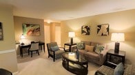 This single-floor residence centers around a thoughtfully designed living area that exudes a warm and inviting ambiance. The space is finished with wall-to-wall carpeting in a neutral tone, contributing to a cozy and comfortable atmosphere throughout.

The living room is anchored by a taupe upholstered sofa accented with decorative teal and earth-toned throw pillows, complemented by a patterned accent chair with a coordinating blue pillow. An oval glass-top coffee table with a dark wood frame serves as a stylish focal point, while a matching end table supports a classic table lamp that casts a soft, warm glow across the room.

The walls are painted in a soft golden-beige hue, providing a neutral backdrop that enhances the carefully curated artwork, including a vibrant abstract canvas and botanical prints. A small dining or accent table with dark chairs occupies a secondary corner, adding functional versatility to the space.

A hallway visible in the background leads to additional areas of the residence, including what appears to be a closet with bi-fold doors. The overall design balances comfort and elegance with a cohesive warm color palette throughout.