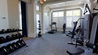 This fitness center offers a bright, welcoming environment designed to support a well-rounded workout routine. The space is finished with durable, low-pile carpet flooring in a neutral gray tone, providing both comfort and practicality underfoot. Large windows dressed with classic plantation shutters line the walls, allowing generous natural light to fill the room while offering adjustable privacy. Elegant arched doorways and architectural details give the space a refined, upscale character that sets it apart from a standard gym setting. Recessed lighting in the ceiling supplements natural illumination, ensuring a consistently well-lit environment at any time of day. The facility is outfitted with a selection of cardio equipment, including a treadmill and a rowing machine, positioned to take advantage of the natural light from the windows. A medicine ball rack stocked with a full range of weighted balls supports functional and strength training needs. Additional strength equipment is visible throughout the space, catering to a variety of fitness goals. Navy blue curtain panels frame the windows, adding a polished, residential aesthetic to the overall design. The combination of thoughtful architectural details, ample natural light, and comprehensive equipment makes this fitness center a practical and inviting space for daily exercise.
