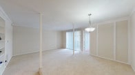 This single-floor residence is designed with a neutral, light-filled aesthetic throughout. The main living area is generously proportioned, featuring soft carpeting, crown molding, and structural columns that add architectural character. A pendant light fixture anchors the dining space, while floor-to-ceiling vertical blinds cover sliding glass doors that open to an outdoor area, allowing abundant natural light to fill the room. Built-in shelving provides practical display and storage space along one wall.

The kitchen offers a functional U-shaped layout with warm hardwood flooring, white cabinetry, speckled countertops, a double-basin sink, and a dishwasher. Ample upper and lower cabinet storage makes this an efficient workspace.

The primary bedroom is spacious, with plush carpeting, a ceiling fan, a natural light-admitting window, and mirrored sliding closet doors that visually expand the room. A dedicated vanity area features a lit mirror, built-in countertop with drawers, and adjacent closet space—an especially practical detail. A second bedroom also includes carpet and a ceiling fan.

The bathroom features a granite-style countertop with an undermount sink, a soaking tub, and a conveniently integrated stacked washer and dryer unit, streamlining laundry within the residence.