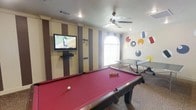 Located at 1800 Links Boulevard in Tuscaloosa, Alabama, this single-story game room is designed as a lively recreational space. The room centers around a full-size pool table with a bold red felt surface, complemented by a ping-pong table positioned nearby, making it well-suited for social and competitive play. A wall-mounted flat-screen television is installed on one wall, supported by a media console, and is framed by vertically striped wall panels in cream, tan, and brown tones that add a structured visual element to the space. The opposite wall features a whimsical mural of colorful circles and geometric shapes, evoking a playful, energetic atmosphere. An arched window allows natural light to filter into the room, brightening the interior. A ceiling fan is centrally mounted, providing air circulation throughout. The flooring features a patterned carpet that ties the room's design elements together. Overall, the space is thoughtfully arranged to maximize usability and entertainment value, offering a welcoming environment for recreation and leisure activities.