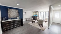 This single-floor business center is thoughtfully designed to balance professional functionality with a welcoming, stylish ambiance. The main area is anchored by a bold navy blue accent wall that serves as a striking focal point, complemented by neutral gray walls, crown molding, and durable wood-look flooring throughout.

The lounge area features an arrangement of upholstered cream chairs surrounding a round wooden coffee table, all set atop a patterned area rug, creating an inviting space for informal meetings or quiet relaxation. A ceiling fan with sleek wooden blades adds comfort overhead, while recessed lighting ensures the space is well-lit throughout.

A dedicated computer workstation along one wall is equipped with dual monitors and pendant lighting overhead, providing a practical workspace for residents or guests. Large windows dressed with layered curtains allow ample natural light to fill the room while maintaining privacy. A wall-mounted flat-screen TV and a dark wood credenza along the navy accent wall add to the room's polished, functional design.

A separate compact kitchenette offers white cabinetry, a stainless steel sink with granite-style countertops, a full-size refrigerator, and a microwave — providing convenient refreshment options. Additional storage and privacy are provided by multiple interior doors accessible from the main space.