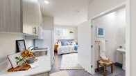 This thoughtfully designed studio unit offers a bright and functional single-floor layout that makes excellent use of every square foot. The open sleeping area serves as the heart of the unit, featuring large windows on multiple walls that invite abundant natural light and provide pleasant outdoor views of surrounding greenery. The warm, neutral color palette and wood-toned flooring throughout create a cohesive and calming ambiance.

The kitchenette is efficiently arranged along one wall, complete with light wood-grain cabinetry, a stainless steel sink, white countertops, and a full-size refrigerator. Upper cabinets provide ample storage while maintaining an open, airy feel. Adjacent to the kitchenette, the entry area offers additional space with room for a small desk or shelving unit, making the most of the layout.

The private bathroom is accessible directly from the main living area and features grey wood-look vinyl flooring, a pedestal sink, a mirrored medicine cabinet, and a toilet. A towel bar and organizational racks keep essentials neatly within reach. The clean white walls and minimalist fixtures give the bathroom a tidy, functional aesthetic. Overall, this studio combines modern finishes, smart storage solutions, and natural light to deliver a comfortable and efficient living environment.