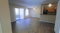 This single-floor space offers a clean, functional layout designed for comfortable everyday living. The main living area is a standout feature, showcasing rich, dark hardwood-style flooring that runs throughout and adds warmth to the interior. A ceiling fan is centrally positioned overhead, providing both practical airflow and a subtle decorative touch beneath a textured ceiling.

Natural light fills the living area generously, courtesy of a large sliding glass door fitted with vertical blinds that opens to an outdoor balcony — ideal for enjoying fresh air and exterior views. An additional window further brightens the space, enhancing the open and airy atmosphere.

Adjacent to the living area, a kitchen is partially visible and features dark countertops, cabinetry in a warm wood tone, and black appliances, offering a practical and cohesive cooking space. A half-wall or bar-style partition creates a natural separation between the kitchen and living areas while maintaining an open-concept feel.

The neutral wall tones throughout complement the flooring and contribute to a versatile backdrop that suits a variety of interior styles. Clean white baseboards and trim provide a crisp finish, rounding out the well-maintained appearance of this inviting single-floor space.