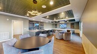 Located at 18183 Wheeler Rd in Westfield, Indiana, this single-floor community gathering space is a thoughtfully designed clubroom that blends upscale finishes with a welcoming, social atmosphere. The space is defined by striking embossed tin-tile ceilings with coffered detailing, complemented by warm hardwood-style flooring and classic white wainscoting along the walls.

The kitchen area serves as a central hub, featuring an expansive granite-topped island with bar seating, a herringbone tile backsplash, open brass-accented shelving, stainless steel appliances, and an embedded television — ideal for entertaining. Adjacent to the kitchen, a lounge and dining zone offers flexible seating arrangements, including a round dining table with upholstered chairs and a modern multi-ring pendant light fixture.

The living area is furnished with tufted leather club chairs arranged around a flat-screen television, creating an inviting media lounge. A billiards table anchors another section of the room, adding a recreational focal point. Large windows and glass French doors flood the interior with natural light and provide views of and access to an outdoor patio area. The overall ambiance is polished yet comfortable, making this a versatile space suited for social gatherings and everyday community use.